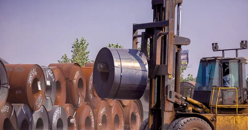 Forklift carrying a hot-rolled steel coil past stacked coil inventory at an Advance Steel yard