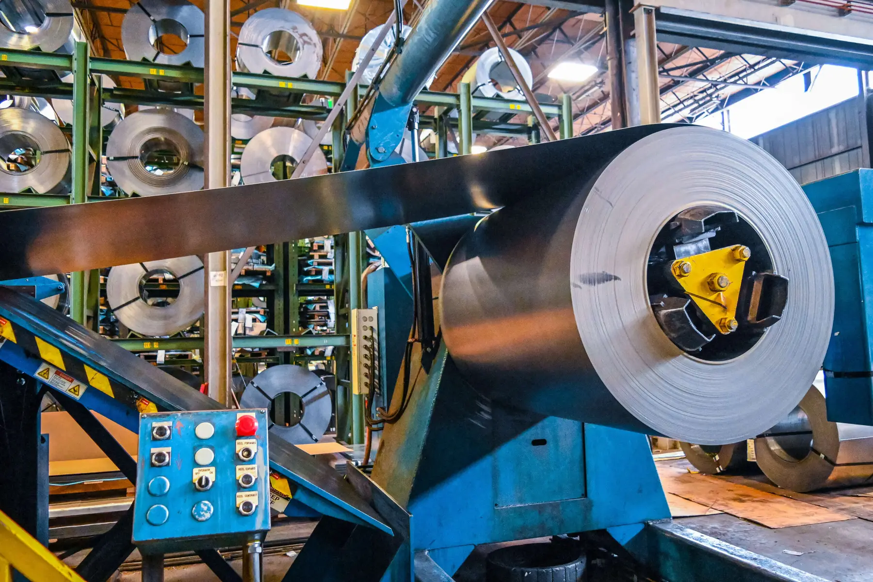 Steel coil mounted on an uncoiler with racks of slit coils behind it
