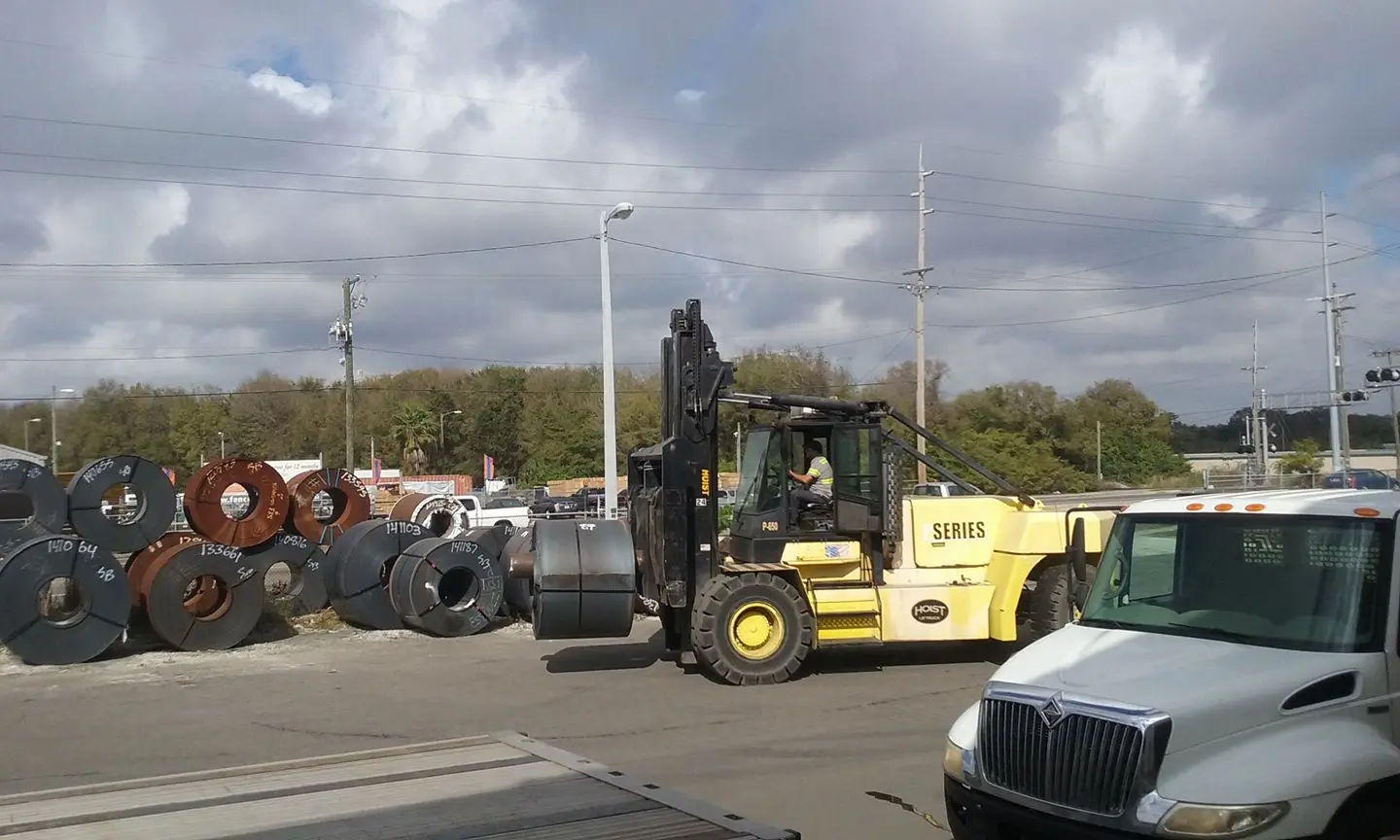 Large forklift moving a steel coil across the Metal Processors yard in Tampa beside rows of coils
