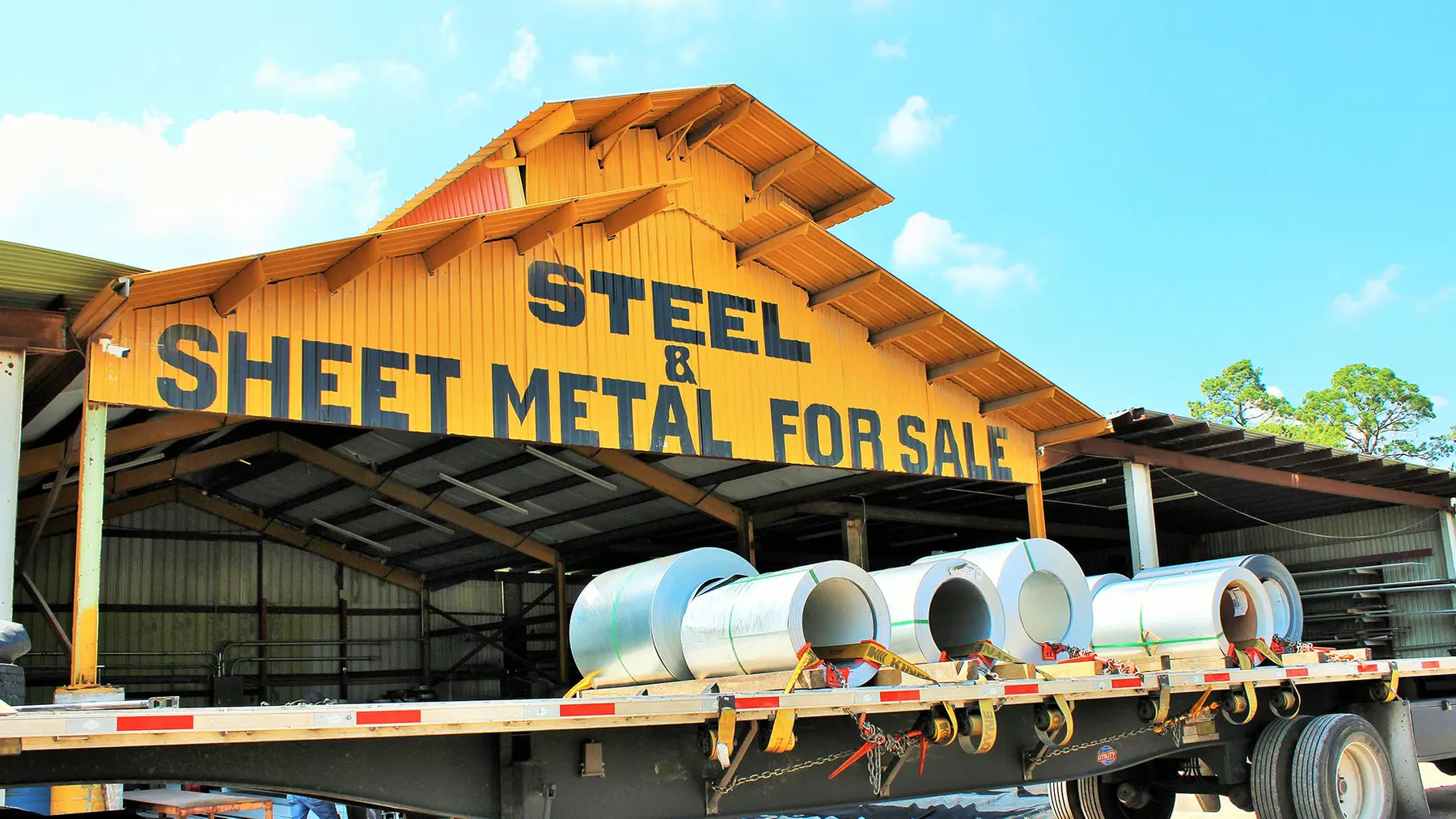 Steel Surplus yard shed with a Steel & Sheet Metal For Sale sign and coils loaded on a flatbed