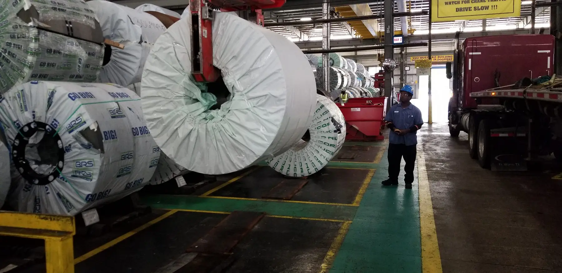 Wrapped master coils staged beside a truck loading bay inside the plant